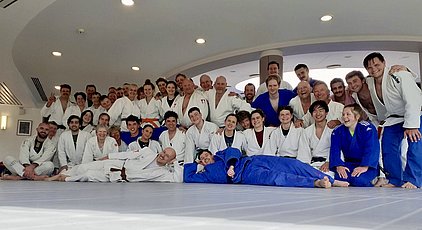 Eine große Gruppe von BJJ-Praktizierenden in weißen und blauen Gi-Anzügen posiert fröhlich auf einer Matte. Die Frauen und Männer stehen und knien zusammen, einige liegen entspannt. Die Atmosphäre wirkt freundlich und einladend, als ob sie gerade ein gemeinsames Training oder Event beendet hätten.