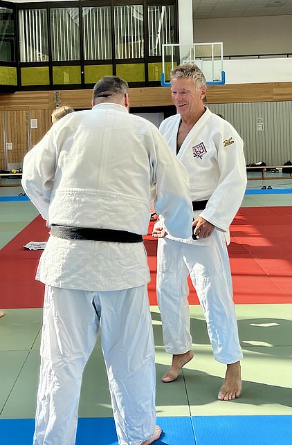 Zwei Judoka in weißen Gis stehen im Training. Der Judoka mit schwarzem Gürtel zeigt ein erleichtertes Lächeln. Im Hintergrund sind Trainingsmatten und die Sporthalle sichtbar.