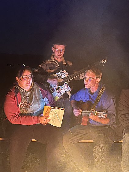 Drei Personen sitzen um ein Lagerfeuer. Eine Frau liest in einem Buch, während zwei Männer Gitarre spielen. Alle wirken entspannt und genießen die Atmosphäre eines nächtlichen Outdoor-Treffens. Das Licht des Feuers beleuchtet die Gesichter und schafft eine gemütliche Stimmung.