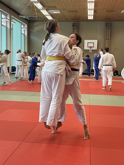 Zwei Frauen in Judokleidung praktizieren einen Wurf im Training. Eine der Frauen hebt die andere an, während im Hintergrund weitere Judokas verschiedene Techniken üben. Die Matten sind rot und grün, und der Raum hat große Fenster mit Licht.