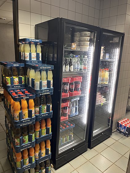 In einem Verkaufsraum stehen zwei gekühlte Getränke-Displays nebeneinander. Links ist ein hohes Regal mit Flaschen verschiedener Säfte und Limonaden, rechts sind zwei Kühlschränke mit Wasser, Softdrinks und weiteren Produkten. Der Boden ist gefliest.