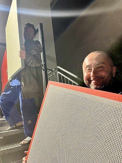 Zwei Männer stehen auf einer Treppe und halten große Platten. Der Mann im Vordergrund lächelt, während der andere im Hintergrund ebenfalls freundlich aussieht. Es ist Nacht, und die Umgebung ist schwach beleuchtet.
