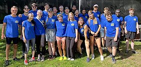 Eine Gruppe von 23 Menschen in blauen T-Shirts steht lächelnd im Freien. Einige tragen Stirnbänder. Die Atmosphäre ist freundlich und entspannt, es scheint sich um ein Gruppenevent oder Teamtreffen zu handeln.