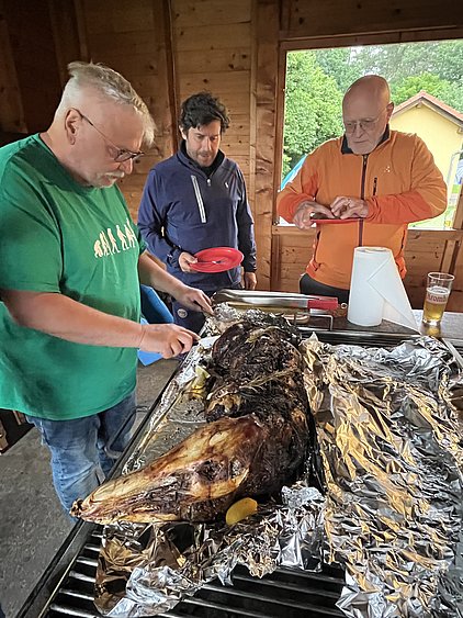 Drei Personen stehen an einem Tisch in einer Holzhütte und bereiten ein gegrilltes Tier vor, das in Alufolie eingewickelt ist. Es sind ein Mann in einem grünen T-Shirt, ein Mann mit dunklem Haar und ein Mann in einem orangefarbenen Jacket.