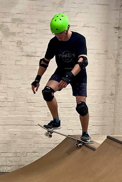 Ein Skateboardfahrer in einem Skatepark balanciert auf einem halbrunden Rampenabschnitt. Er trägt einen grünen Helm und Schutzausrüstung an Ellenbogen und Knien. Der Fahrer ist in Bewegung und zeigt eine dynamische Pose. Der Hintergrund ist eine weiße Ziegelwand.