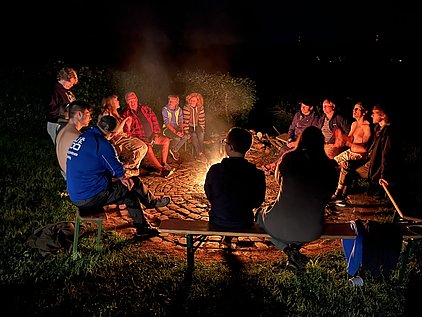 Eine Gruppe von Menschen sitzt um ein Lagerfeuer in der Nacht. Sie unterhalten sich und lachen. Die Atmosphäre ist entspannt und gesellig, mit der warmen Flamme des Feuers im Mittelpunkt. Es ist eine gesellige Zusammenkunft in der Natur.