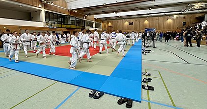 Eine Gruppe von Judokas in weißen Uniformen trainiert auf matten in verschiedenen Farben. Im Vordergrund sind mehrere Paar Sandalen zu sehen. Zuschauer sitzen auf Bänken im Hintergrund, beobachtend das Training.