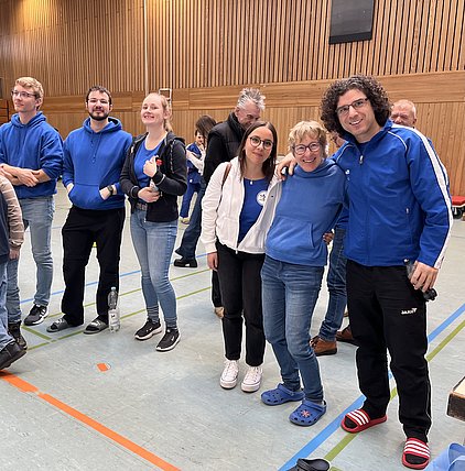 Eine Gruppe von sechs Personen steht in einer Sporthalle. Sie tragen blau-weiße Kleidung und lächeln freundlich in die Kamera. Im Hintergrund sind weitere Personen zu sehen. Die Atmosphäre wirkt entspannt und freundlich, als ob sie an einem gemeinsamen Event teilnehmen.
