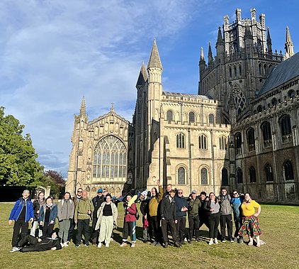 Eine Gruppe von 20 Personen posiert fröhlich vor einer beeindruckenden Kathedrale mit gotischer Architektur. Im Hintergrund sind hohe Türme und bunte Fenster sichtbar. Der Himmel ist blau mit einigen Wolken, und die Menschen tragen verschiedene farbige Kleidungsstücke.