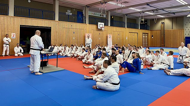 Eine Gruppe von Judokas in weißen und blauen Anzügen sitzt auf einer Judomatte. Ein Trainer in einem weißen Anzug steht an einem Tisch mit einem Laptop und spricht zu den Teilnehmern, die aufmerksam zuhören. Im Hintergrund sind weitere Judokas und eine Sporthalle sichtbar.