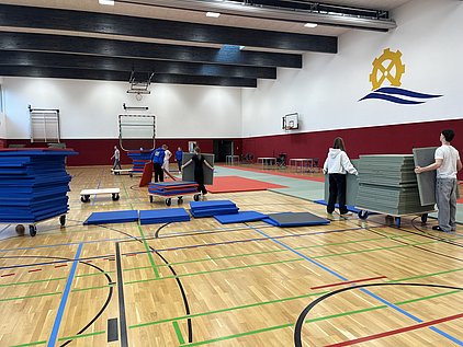 In einer Sporthalle helfen mehrere Personen beim Aufstellen von Sportmatten. Einige tragen Matten auf Rollwagen, während andere sie ordentlich stapeln. Der Fußboden ist aus Holz mit bunten Linien, und im Hintergrund sind Basketballkörbe sichtbar.