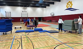 In einer Sporthalle helfen mehrere Personen beim Aufstellen von Sportmatten. Einige tragen Matten auf Rollwagen, während andere sie ordentlich stapeln. Der Fußboden ist aus Holz mit bunten Linien, und im Hintergrund sind Basketballkörbe sichtbar.