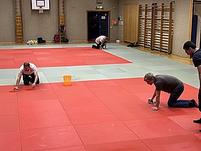 In einer Turnhalle reinigen mehrere Personen die Matten. Einige knien auf dem Boden und wischen die rote Fläche mit Tüchern ab. Ein gelbes Eimerchen steht in der Nähe. Im Hintergrund sieht man die Wände der Halle und eine Basketballkorbanlage.