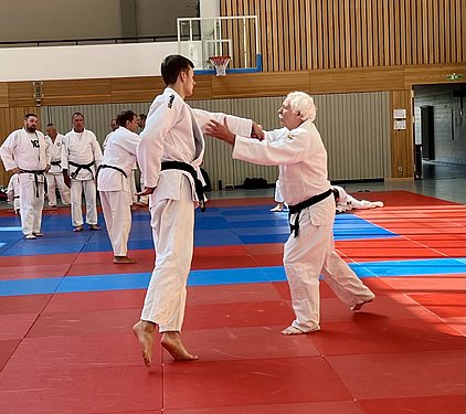 Eine Gruppe von Judoka trainiert auf einer Matte. Im Vordergrund stehen zwei Kämpfer, einer in einem weißen Gi mit schwarzem Gürtel, der andere in einem grauen Gi. Der Trainer zeigt eine Technik, während andere Judoka im Hintergrund üben oder zuschauen.