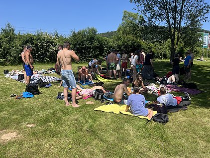 Eine Gruppe von Menschen entspannt sich auf einer Wiese. Einige sitzen oder liegen auf Handtüchern, während andere stehen und miteinander sprechen. Es ist sonnig und grün, was eine sommerliche Atmosphäre schafft.