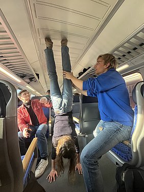 Drei junge Menschen sitzen in einem Zug. Eine Person steht auf den Händen, während zwei andere sie stützen. Die Szene vermittelt eine lockere, unerwartete Atmosphäre und zeigt, wie die Gruppe Spaß hat.