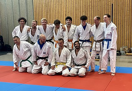 Eine Gruppe von zwölf Männern inJudoanzügen steht und sitzt zusammen auf einer farbigen Matte. Einige tragen unterschiedliche Gürtel in verschiedenen Farben, darunter Schwarz, Blau, Grün und Gelb. Sie lächeln und posieren freundlich für das Foto in einer Sporthalle mit hölzernen Wänden.