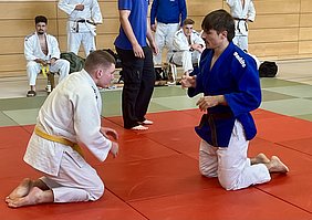 Zwei Judokas befinden sich auf einer roten Matte. Der Judo-Kämpfer in einem weißen Gi mit gelbem Gürtel kniet, während der Kämpfer in einem blauen Gi mit braunem Gürtel ihm gegenübersteht und eine Technik demonstriert. Im Hintergrund sind weitere Judokas zu sehen, die zuschauen.