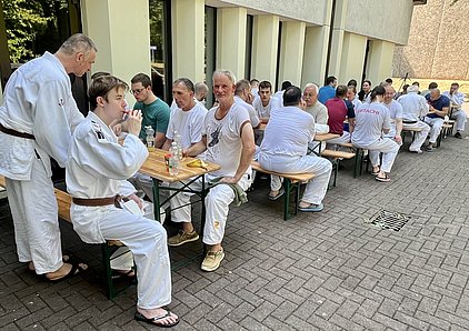 Eine Gruppe von Menschen in weißen Trainingsanzügen sitzt an langen Tischen im Freien. Einige unterhalten sich, während andere essen oder trinken. Die Szenerie vermittelt eine gesellige Atmosphäre nach einem Sporttraining.