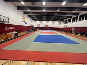 Das Bild zeigt eine Judo-Halle mit einem großen, farbigen Mattenbereich. Die Matten sind rot, blau und grün angeordnet. Am Rand stehen Stühle und ein Tisch mit Informationen zum Hessischen Judo Verband. Im Hintergrund sind Basketballkörbe und große Fenster sichtbar.