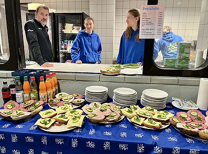 In der Abbildung sind eine Reihe belegter Brote mit verschiedenen Belägen wie Wurst, Käse und Gurkenscheiben auf einem Tisch zu sehen. Im Hintergrund stehen zwei Personen in blauen Pullovern an einer Theke neben einer Preisliste für Getränke und Snacks.