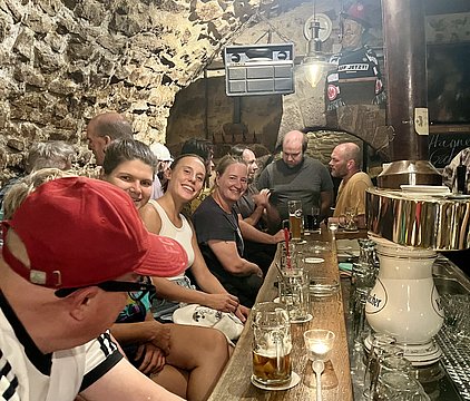 Eine Gruppe von Menschen sitzt an einer langen Theke in einem urigen Keller. Sie lächeln, unterhalten sich und genießen Getränke. Im Hintergrund ist eine Steinwand sichtbar, die für die Atmosphäre des Ortes sorgt. Ein Getränkespender steht auf der Theke.