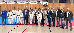Eine Gruppe von Judokas und Offiziellen steht auf einer Mattenfläche in einer Sporthalle. Sie tragen Judogis in verschiedenen Farben. Im Hintergrund ist ein Banner zu sehen, das den Sportverband begrüßt. Die Stimmung ist freundlich und feierlich.