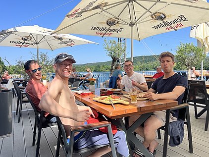 Vier Männer sitzen an einem Tisch im Freien, umgeben von Sonnenschirmen. Sie lächeln und halten Essen in den Händen. Hinter ihnen ist eine Wasserfläche und Bäume zu sehen, was auf eine entspannte Atmosphäre hinweist.