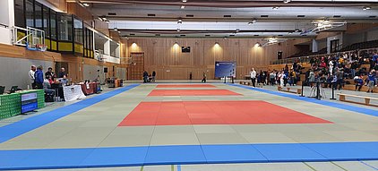 Eine Sporthalle mit Matten in verschiedenen Farben: einige rote und andere grüne Flächen. Auf der linken Seite stehen Tische mit technischen Geräten, während sich Zuschauer auf den Tribünen versammeln. Im Hintergrund sind Menschen, die sich auf ein Wettkampfereignis vorbereiten.