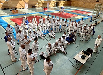 Eine Gruppe von Judoka in weißen Gi stehen und sitzen in einer Sporthalle. Im Vordergrund spricht ein Trainer zu den versammelten Athleten, während ein paar Teilnehmer Notizen machen. Die Halle ist mit Matten in verschiedenen Farben ausgelegt.