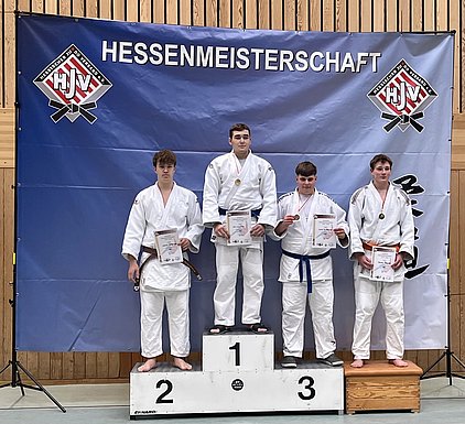 Vier Judokas stehen auf einem Siegerpodium. Der erste Platz trägt eine Goldmedaille, der zweite Platz eine Silbermedaille, und der dritte Platz zeigt eine Bronzemedaille. Alle halten Urkunden in ihren Händen. Im Hintergrund ist ein Banner mit der Aufschrift "Hessenmeisterschaft" zu sehen.