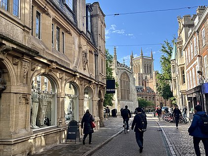 Eine geschäftige Straße mit historischen Gebäuden, darunter ein Geschäft mit dem Namen „REISS“. Menschen gehen, fahren Rad und stehen auf dem Gehweg. Im Hintergrund sind hohe Kirchtürme und grünes Laub sichtbar, unter einem klaren blauen Himmel.