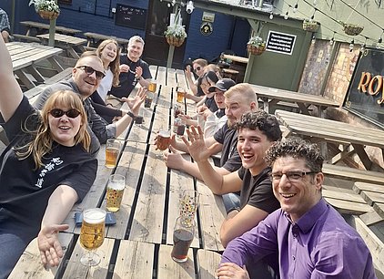 Eine Gruppe von Freunden sitzt an einer langen Holztabelle im Freien, die mit verschiedenen Getränken gefüllt ist. Viele lachen und winken in die Kamera. Die Atmosphäre wirkt fröhlich und entspannt, es ist sonnig. Im Hintergrund sind weitere Tische und Pflanzen zu sehen.