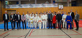 Eine Gruppe von Judoka und Gästen steht auf einer Judomatte in einer Halle. Die Judoka tragen weiße und orange Gürtel. Im Hintergrund sind Banner mit dem Logo der Judoschule und Informationen über das Training sichtbar. Die Atmosphäre ist freundlich und gemeinschaftlich.