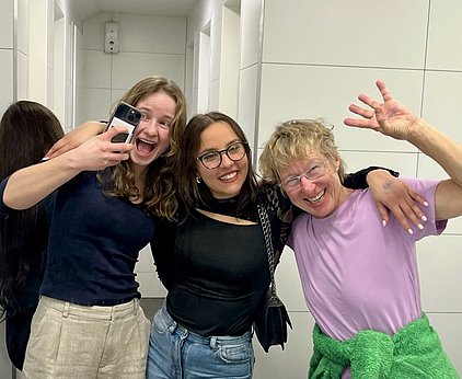 Drei Frauen stehen in einem Bad und lächeln fröhlich in die Kamera. Eine Frau hält ein Handy und macht ein Selfie. Die anderen beiden umarmen sie. Die Stimmung ist ausgelassen und freundlich. Die Wände sind hell und modern gestaltet.
