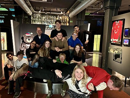 Eine Gruppe von zwölf Menschen posiert in einem Museum. Einige sitzen auf Stufen, andere stehen hinter ihnen. Zwischen ihnen steht ein Stoffbär mit einer Weihnachtsmütze. Im Hintergrund sind Ausstellungsstücke wie ein rotes Trikot und Pokale sichtbar. Alle wirken fröhlich und lachen.