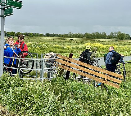 Eine Gruppe von vier Personen steht neben ihren Fahrrädern an einem Feldweg. Zwei Personen heben ein Fahrrad über ein Zaungitter. Im Hintergrund sind Wiesen und Bäume zu sehen, der Himmel ist bewölkt. Schilder weisen auf einen öffentlichen Fußweg hin.