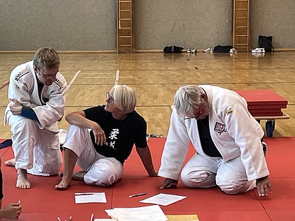 In einem Sportraum sitzen drei Personen, die Judoanzüge tragen. Sie diskutieren zusammen auf einer roten Matte und haben einige Unterlagen vor sich. Die Atmosphäre wirkt kollegial und lehrreich, während sie sich auf eine Trainingseinheit vorbereiten.