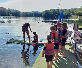 Ein Erwachsener steht auf einem Paddleboard und hilft einem Kind, während eine Gruppe von mehreren Kindern in Schwimmwesten am Ufer zuschaut. Der Hintergrund zeigt einen ruhigen See und Bäume. Es ist ein sonniger Tag.