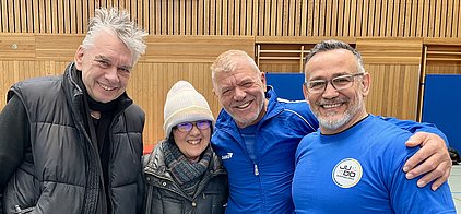 Vier Personen lächeln für die Kamera in einer Sportumgebung. Sie tragen sportliche Kleidung, darunter ein blaues Judo-Shirt. Die Wand hinter ihnen ist aus Holz, und es gibt Trainingsgeräte im Hintergrund. Die Gruppe strahlt Freude und Zusammenhalt aus.