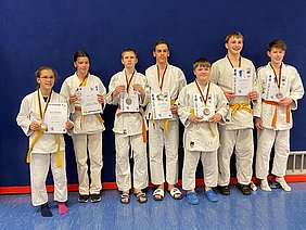 Eine Gruppe von sieben jungen Judoka in weißen Judogatters, alle tragen Medaillen und halten Urkunden in den Händen. Sie lächeln stolz in die Kamera. Im Hintergrund ist eine blaue Wand sichtbar.