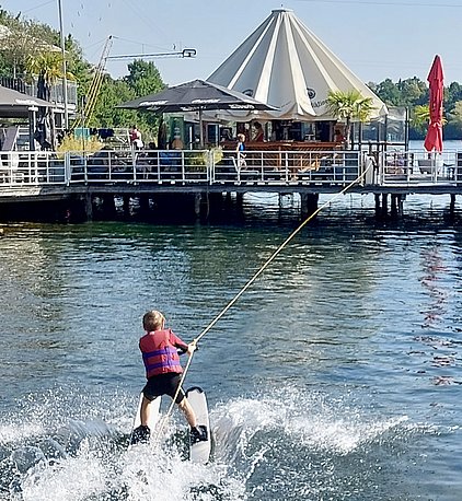 Ein Junge mit einer Schwimmweste fährt auf einem Wasserboard und wird von einem Seil gezogen. Er gleitet über die Wasseroberfläche, während Spritzer hinter ihm aufsteigen. Im Hintergrund ist ein Café an einem Steg sichtbar. Es ist sonnig und die Umgebung wirkt lebhaft.