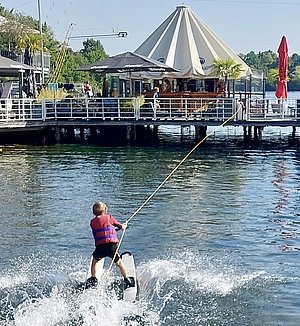 Ein Junge mit einer Schwimmweste fährt auf einem Wasserboard und wird von einem Seil gezogen. Er gleitet über die Wasseroberfläche, während Spritzer hinter ihm aufsteigen. Im Hintergrund ist ein Café an einem Steg sichtbar. Es ist sonnig und die Umgebung wirkt lebhaft.