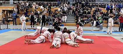 Eine Gruppe von Kindern in Judokleidung trainiert auf einer roten Matte. Einige Kinder liegen auf dem Boden, während andere sich auf das Training konzentrieren. Im Hintergrund sind Zuschauer auf den Tribünen zu sehen, die das Geschehen beobachten.