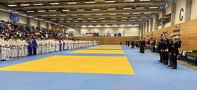 Das Bild zeigt eine Judoveranstaltung in einer Halle. Auf der linken Seite stehen Teilnehmer in weißen Judokis, während rechts Trainer und Offizielle in Anzügen aufgestellt sind. In der Mitte befindet sich eine Judomatte in Blau und Gelb, umgeben von einem Publikum auf Tribünen.