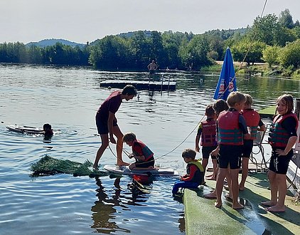 Eine Gruppe von Kindern in Rettungswesten spielt am Ufer eines Gewässers. Einige Kinder stehen auf einer Plattform, während andere im Wasser schwimmen. Ein Erwachsener hilft einem Kind auf einem Brett. Im Hintergrund sind Bäume und ein Steg sichtbar.