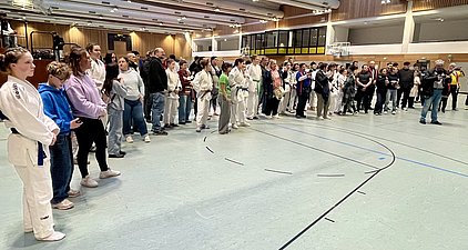 Eine Gruppe von Menschen steht in einer Sporthalle. Einige tragen Judogi (Judoanzüge), während andere in Alltagskleidung gekleidet sind. Die Teilnehmer scheinen auf eine Veranstaltung oder einen Wettkampf zu warten. Es sind auch Zuschauer im Hintergrund zu sehen.