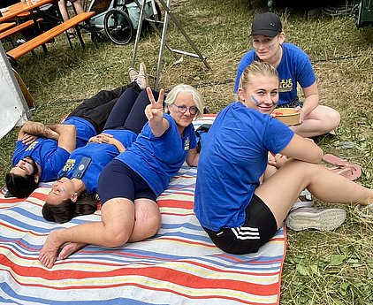 Fünf Personen sitzen und liegen entspannt auf einer gestreiften Decke im Freien. Einige tragen blaue T-Shirts. Eine Frau mit grauen Haaren zeigt ein Peace-Zeichen. Die Atmosphäre wirkt freundlich und ausgelassen.
