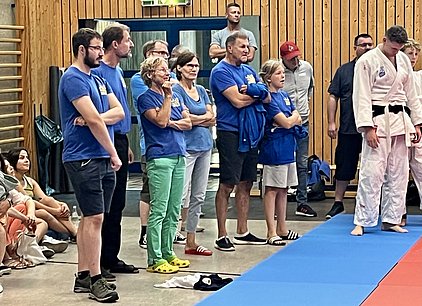 Eine Gruppe von Menschen in blauen T-Shirts beobachtet einen Judo-Wettkampf. Die Zuschauer stehen am Rand der Matte, einige sind gespannt, andere zeigen Interesse. Im Vordergrund steht ein Judo-Kämpfer in weißem Gi mit einem schwarzen Gürtel.