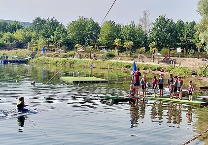Eine Gruppe von Kindern steht an einem Steg am Wasser, während ein Jugendlicher mit einem Seil hinter einem Boot fährt. Alle Kinder tragen Schwimmwesten. Im Hintergrund sind Bäume und Freizeiteinrichtungen sichtbar. Es ist ein sonniger Tag.